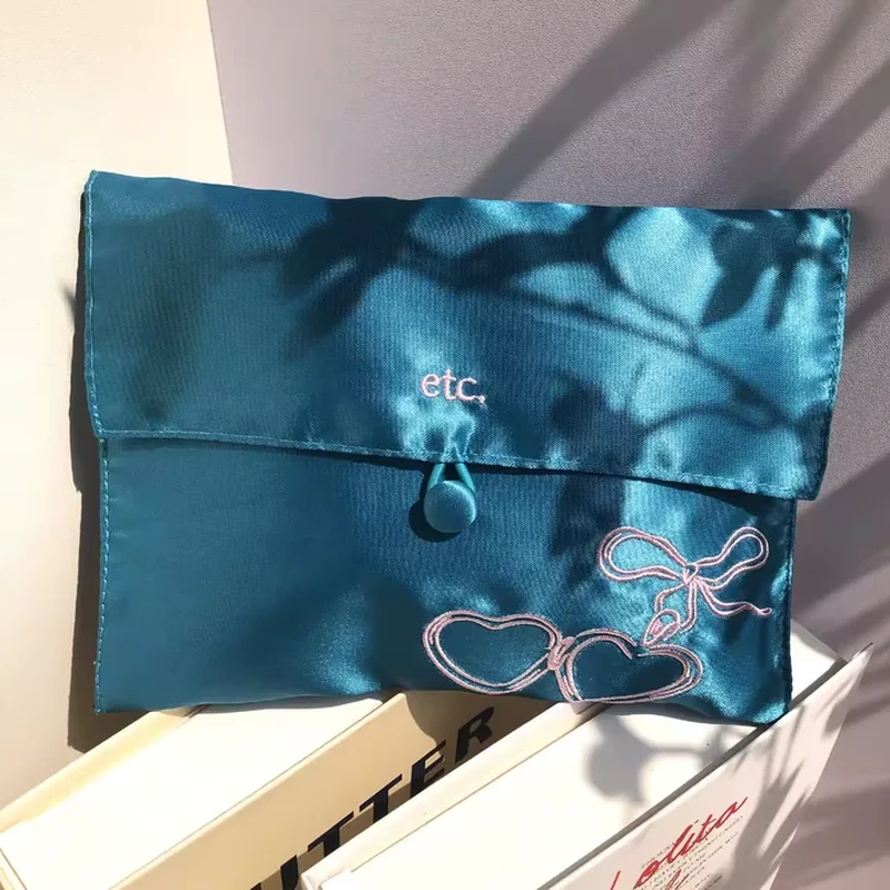 A fabric-covered button and short loop close a broad satin envelope with corner embroidery