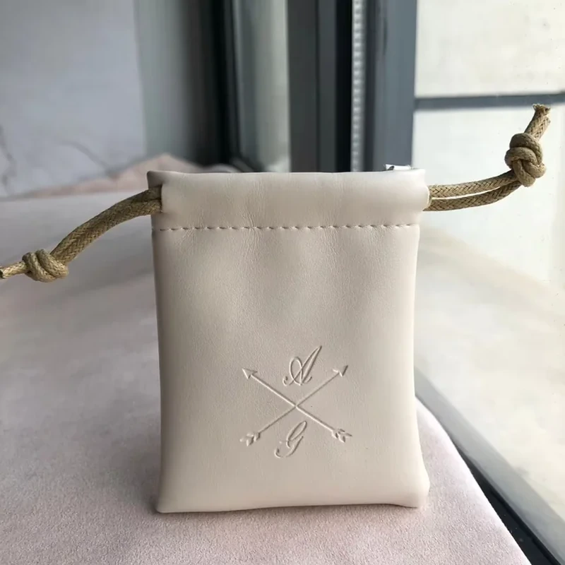 Cream square leather-look pouch with embossed crossed-arrow emblem and tan cord