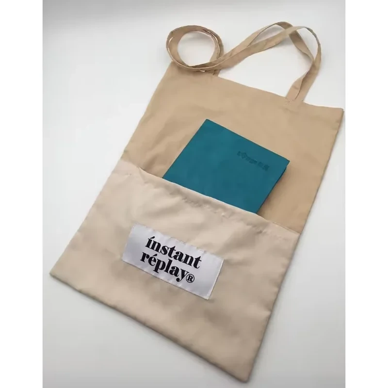 A pale long handle tote has a large outer pocket holding a notebook and a small label
