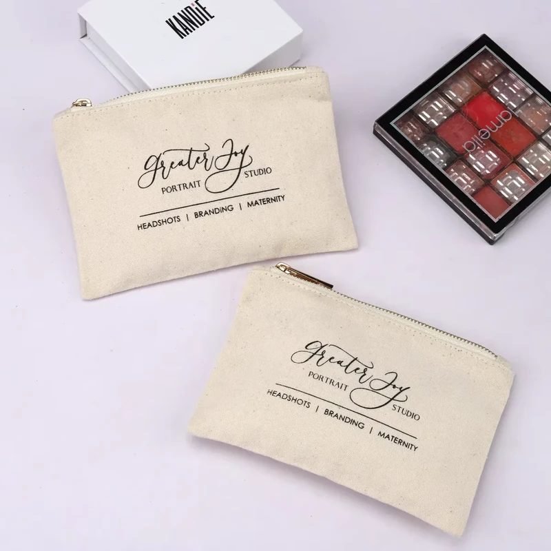 Two cream makeup pouches with printed script logos and gold-tone zippers.