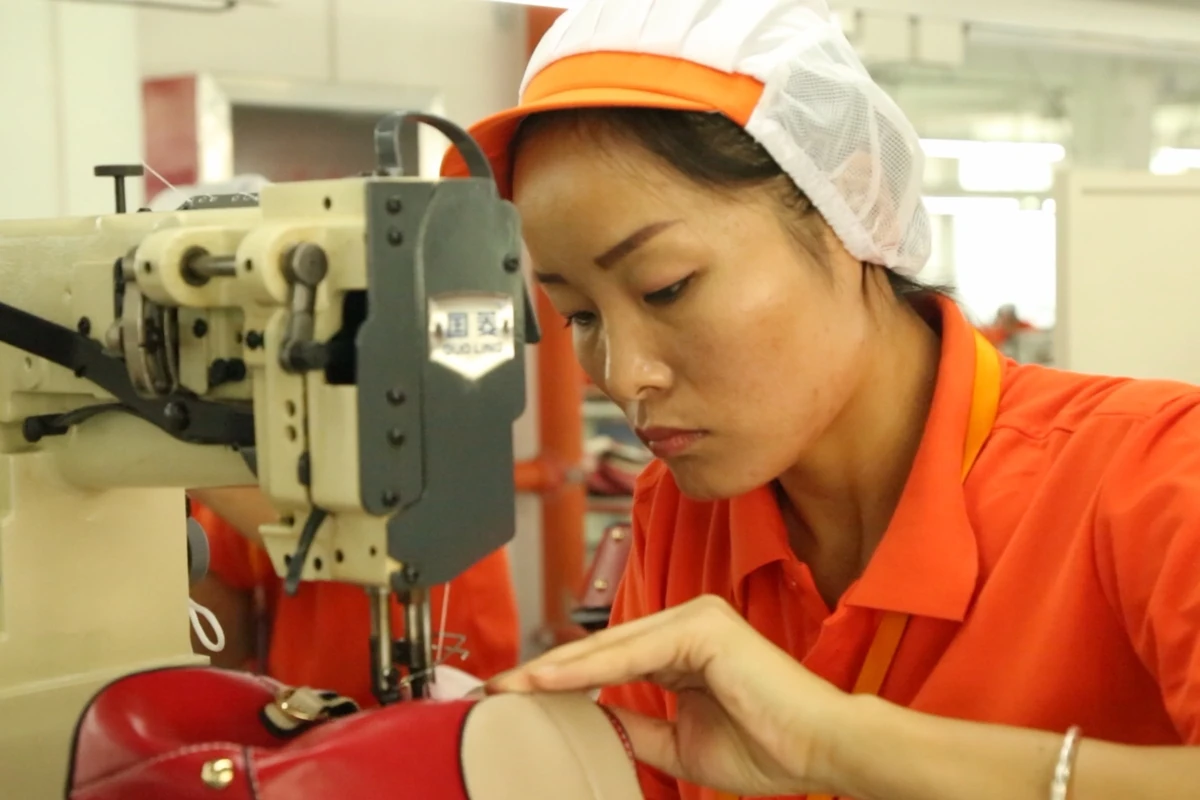 An operator stitching the edge of a red bag at an industrial sewing machine.
